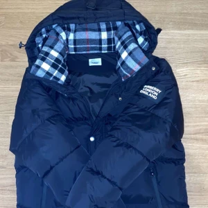 Svart pufferjacka från Burberry - Svart pufferjacka från Burberry med avtagbara ärmar, så du kan bära den som väst. Jackan har huva och insida med klassiskt rutigt foder i grått, vitt och rött. Märkespatch med 'Burberry London England' på bröstet. Två dragkedjefickor framtill.