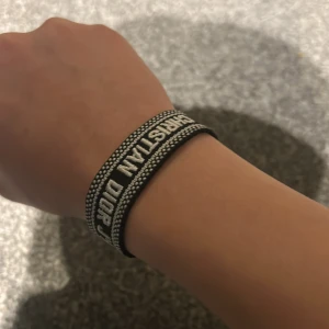 Svart armband Christian Dior J'Adior - Snyggt svart armband från Christian Dior med texten 'J'Adior' broderad i vitt. Armbandet har justerbar knytning med tofsar och detaljerade kanter. Perfekt accessoar för att lyfta din outfit med en lyxig touch.