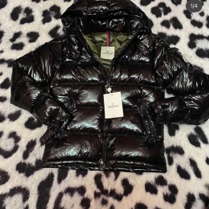 Svart pufferjacka från Moncler - Säljer en glansig svart pufferjacka från Moncler med huva och dragkedja framtill. Jackan har quiltad design och långa ärmar med snygga detaljer vid mudden. Perfekt för kalla dagar och riktigt snygg streetstil.