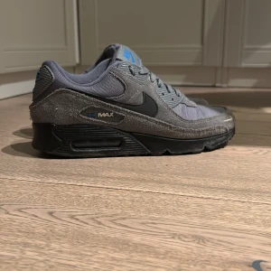 Nike Air Max 90 grå sneakers - SKORNA TVÄTTAS INNAN KÖPET!!!!!!!            Tjena!  Säljer ett par grå Nike Air Max 90 sneakers med svart sula och blå detaljer på tungan och loggan. Skorna har en kombination av mesh och mocka på ovandelen, klassisk snörning och rund tå. Perfekt för dig som gillar stilrena och sportiga sneakers samt så håller dom bra höst/vintern.