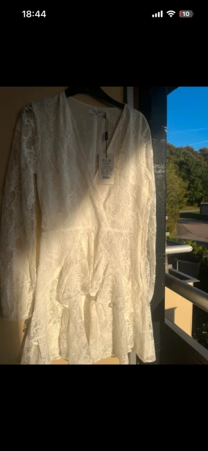 Vit spetsklänning från NA-KD - Supersöt vit kortklänning från NA-KD med v-ringning och långa ärmar. Klänningen har spetsmaterial och volangdetaljer nedtill som ger en romantisk vibe. Perfekt för sommarens alla tillfällen, och den har en snygg omlott-look framtill.