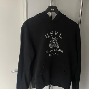 Svart hoodie från Ralph Lauren - Svart hoodie från Ralph Lauren med tryck på bröstet som visar en motorcykel och texten U.S.R.L. samt Ralph Lauren U.S.No. Tröjan har huva, magficka och långa ärmar. Perfekt för dig som gillar streetwear och sportig stil.