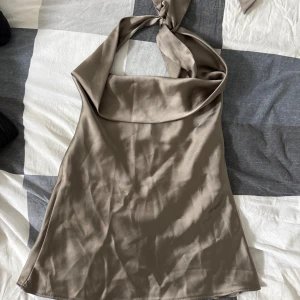Beige omlott halterneck topp - Säljer en beige omlott halterneck topp i satinliknande material. Toppen har en snygg knytning i nacken och öppen rygg, vilket ger en trendig och chic look. Perfekt för dig som gillar stilrena och moderna plagg med lite extra edge.