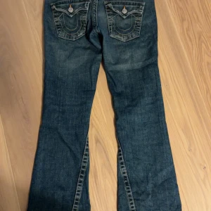 True Religion bootcut jeans blå, stl W28 - Säljer ett par klassiska blå jeans från True Religion med bootcut passform. Jeansen har snygga kontrastsömmar, ikoniska ficklock bak med silverfärgade knappar och tydlig logga. Tillverkade i kraftigt denim med slitna detaljer för en cool look. Lite slitna längst ner men annars bra