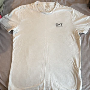 Vit EA7 Emporio Armani t-shirt - Snygg vit t-shirt från EA7 Emporio Armani med diskret logga på bröstet och svart detalj på ryggen. Klassisk rund hals och korta ärmar. Tillverkad i mjuk bomull för en skön känsla. Perfekt för dig som gillar stilrena och exklusiva plagg.