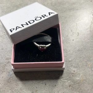 Pandora silverring med röd hjärtformad sten - Säljer en söt silverring från Pandora med en röd hjärtformad sten i mitten. Ringen har en fin, vågig design och levereras i originalask. Perfekt för dig som vill ha något romantiskt och stilrent.