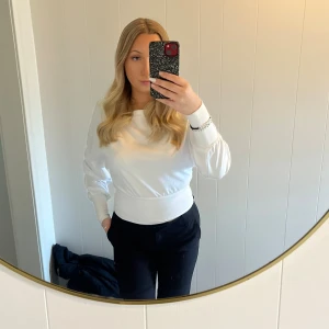 Vit långärmad tröja med bred mudd - Säljer en stilren vit långärmad tröja med bred ribbad mudd i midjan och vid ärmsluten. Tröjan har en rund halsringning och är tillverkad i mjukt bomullsmaterial. Passformen är normal och den har en clean look som är enkel att matcha med olika outfits. Använd endast en gång