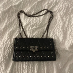 Svart quiltad axelväska med nitar - Snygg svart axelväska med quiltat mönster och silverfärgade nitar. Väskan har en kedjerem i metall och ett stilrent lås framtill. Perfekt för dig som gillar en edgy och trendig look.