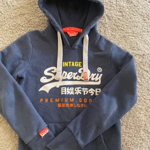 Marinblå Superdry hoodie XS - Snygg marinblå hoodie från Superdry med stor logga och japanska tecken på bröstet. Tröjan har huva med grova vita snören, känguruficka och ribbade muddar. Perfekt för dig som gillar streetwear och vill ha en cool look.