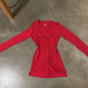 Röd långärmad topp från EGMP - Säljer en röd långärmad topp från EGMP i 100% bomull. Toppen har v-ringning och knappar hela vägen framtill, vilket ger en snygg och enkel look. Passformen är normal och materialet är mjukt och bekvämt. Perfekt för dig som gillar stilrena och färgstarka plagg.