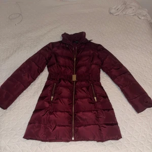 Vinröd pufferjacka från KappAhl - Snygg vinröd pufferjacka från KappAhl med guldiga dragkedjor och bälte i midjan. Jackan har hög krage, långa ärmar och två fickor med dragkedja. Materialet är glansigt och känns mjukt, perfekt för kalla dagar. Stängs med dragkedja framtill.