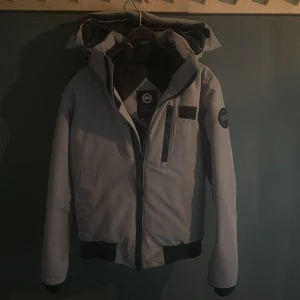 Grå pufferjacka från Canada Goose - Säljer en grå pufferjacka från Canada Goose med huva och svarta detaljer. Jackan har dragkedja framtill, bröstficka med dragkedja och ribbade muddar. Märkespatch på ärmen och stilren design som håller dig varm under kalla dagar. Köptes på Canada goose affär i New York för ca 13 000kr.