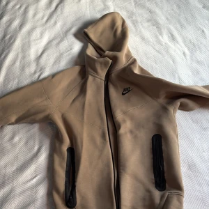 Beige Nike hoodie med svarta detaljer XS - Säljer en beige hoodie från Nike i storlek XS. Tröjan har huva, svarta dragkedjor på fickorna och en svart Nike-logga på bröstet. Materialet är mjukt och känns som bomull och polyester. Perfekt för chill eller träning, snygg och enkel design. Den har ett hål i ryggen som man har sytt för men kan synas när man har på sig plagget, pris kan diskuteras!