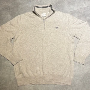 Beige half zip tröja från GANT - Snygg beige tröja från GANT med half zip och ribbad krage med svarta ränder. Tröjan har broderad logga på bröstet och är långärmad med ribbade muddar. Perfekt för dig som gillar klassisk stil och vill ha något som funkar året runt och är inte änvänd