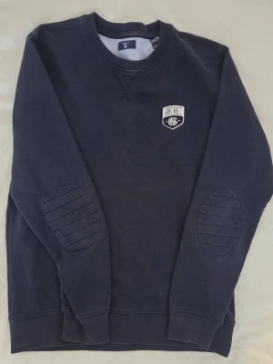 Mörkblå sweatshirt från Gant - Säljer en mörkblå sweatshirt från Gant med broderad logga på bröstet och förstärkta armbågslappar med ribbade detaljer. Tröjan har rund hals och långa ärmar, tillverkad i mjuk bomullsmix som är skön att bära. Perfekt för chill dagar och snygg till jeans.