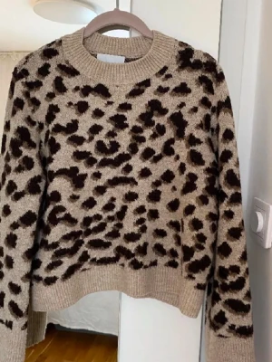 Beige leopardmönstrad stickad tröja - Säljer en beige stickad tröja med leopardmönster i svart och brunt. Tröjan har rund halsringning, ribbade muddar och är långärmad. Perfekt för dig som vill ha en trendig och mysig look under kyliga dagar.