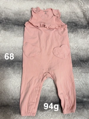 Rosa byxdress med hjärtfickor - Supersöt rosa byxdress från Polarn O. Pyret med volangdetalj vid bröstet och två hjärtformade fickor framtill. Byxdressen har tryckknappar på axlarna och resår vid bensluten för extra komfort. Perfekt för små barn som vill vara både bekväma och gulliga.🌸 Fint använt skick. Storlek 68