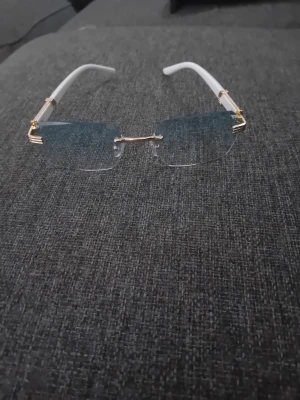 Solglasögon - Säljer ett par stilrena glasögon med blåtonade rektangulära linser och vit båge. Skalmarna har ett snyggt marmormönster i svart och vitt samt detaljer i guld. Perfekt för dig som vill ha en unik och trendig look.