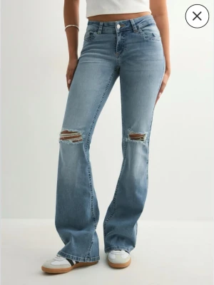 Bootcut ljusblå jeans med slitningar - Säljer ett par ljusblå bootcut Nelly jeans med snygga slitningar på knäna och råa kanter vid bensluten. Baktill har fickorna dekorativa detaljer. Tillverkade i mjukt denimtyg.