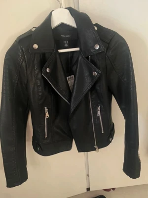 Svart skinnjacka från Vero Moda XS - Säljer en svart bikerinspirerad skinnjacka från Vero Moda i storlek XS. Jackan har silvriga dragkedjor, bred krage med tryckknappar och detaljerade sömmar på ärmarna. Snygg kort modell med coola zip-detaljer vid ärmslut och fickor.