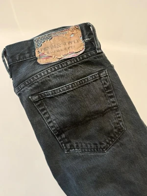 Svarta jeans från Denim & Supply (Ralphlauren jeans) - Svarta jeans från Denim & Supply med klassisk femficksdesign och slitstark denim. Jeansen har rak passform och metallknappar fram. Snyggt sliten etikett baktill med mönsterdetaljer. Perfekta för dig som gillar en enkel och cool stil.