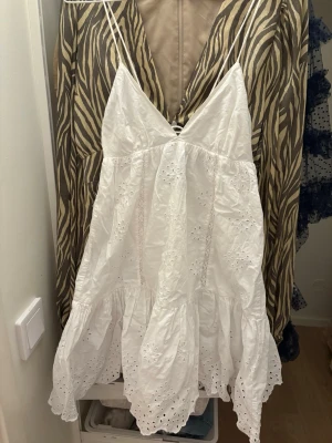 Vit broderad sommarklänning från Zara - Supersöt vit kortklänning från Zara i bomull med tunna axelband, broderade blommönster och volangkant. Klänningen har omlottkänsla baktill med knyt och öppen detalj. Perfekt för varma dagar och midsommar!