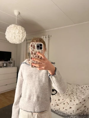 Grå stickad tröja med mörkblå kant - Mysig ljusgrå stickad tröja med lång ärm och cool mörkblå kant längs krage och ärmslut. Tröjan har en loose passform och är perfekt för kyliga dagar. Materialet är mjukt och skönt, och detaljerna ger en unik touch till din outfit.