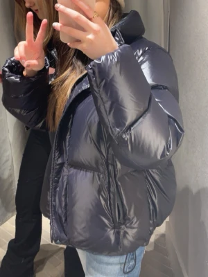 Svart pufferjacka från Gant - Säljer en svart, glansig pufferjacka med hög krage och dragkedja framtill. Jackan är oversized och har en riktigt fluffig look som håller dig varm under kalla dagar. Perfekt för dig som gillar streetstyle och vill ha en jacka som sticker ut.