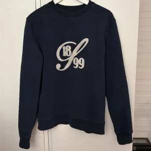 Mörkblå sweatshirt Stenströms M - Mörkblå sweatshirt från Stenströms i storlek M. Tröjan har ett broderat tryck med siffrorna 1899 på bröstet och ribbade muddar vid ärmslut och hals. Klassisk rund halsringning och långärmad modell, perfekt för en clean och stilren look.