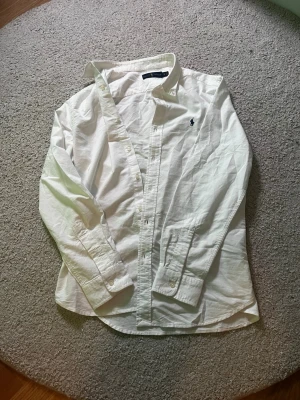 Vit skjorta från Ralph Lauren - Klassisk vit skjorta från Ralph Lauren i bomull med button-down krage och broderad marinblå logga på bröstet. Skjortan har lång ärm, knappar framtill och en tidlös, clean look som passar till allt.