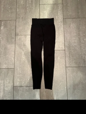 Svarta höga leggings med ribbade detaljer - Säljer ett par svarta leggings med hög midja och bred linning. Byxorna har ribbade detaljer längs benen och är tillverkade i ett stretchigt material som sitter tight. Perfekta för dig som gillar en clean och sportig look.