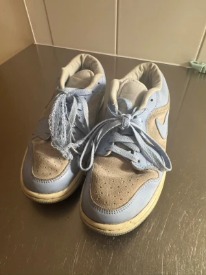 Nike Jordan 1 Low i ljusblå mocka - Säljer ett par Nike Jordan 1 Low i en snygg ljusblå färg med grå mocka detaljer. Dem är använda mycket, vilket har gjort att ena skosnöret är sönder därav behöver man köpa nya. 