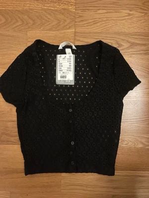 Svart blus med knappar från H&M - Kort svart blus från H&M i återvunnen polyester med broderade hålmönster och knappar framtill. Modellen har korta ärmar, djup rundringning och croppad passform. Perfekt för dig som gillar detaljer och en luftig känsla.