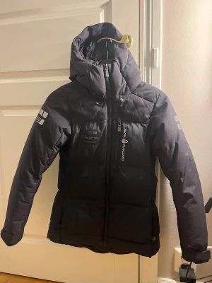 Svart dunjacka Sail Racing med huva - Svart dunjacka från Sail Racing med stor, mysig huva och  loggor på ärm och rygg. Jackan har dragkedja framtill,  och är fylld med Allied Feather Down (500 fill power). Perfekt för kalla dagar och har en clean, sportig look.