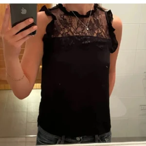 Svart blus med spets och volang - Säljer en svart blus med spets upptill och volangdetaljer vid axlarna. Blusen är ärmlös och har en rund halsringning med dekorativ spets som ger en snygg look. Perfekt att styla med jeans eller kjol för en trendig outfit.