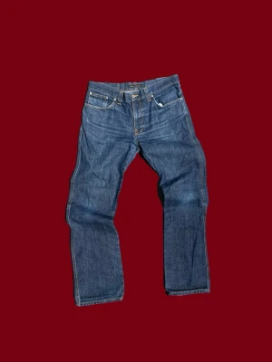 Nudie Jeans Slim Jim blå jeans W34 L32 - Säljer ett par Nudie Jeans Slim Jim i klassisk blå tvätt med snygga orangea sömmar och broderad bakficka. Modellen är rak och slim, tillverkad i 100% bomull. Passar dig som gillar stilrena jeans med schysst passform.