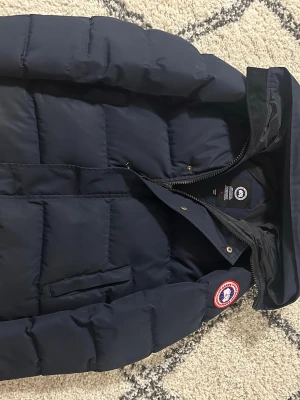 Mörkblå pufferjacka från Canada Goose - Säljer en mörkblå pufferjacka från Canada Goose med klassisk patch på ärmen. Jackan har huva, dragkedja och tryckknappar framtill. Fylld med dun och har en quiltad design som ger extra värme. Perfekt för kalla dagar och snygg till streetwear.