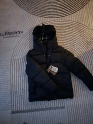 Svart dunväst från Burberry - Svart dunväst från Burberry med avtagbara ärmar och huva. Insidan har klassiskt rutigt Burberry-foder i grått, vitt, svart och rött. Västen har två fickor med dragkedja och tryckknappar framtill. Perfekt för dig som vill ha en mångsidig och snygg look.