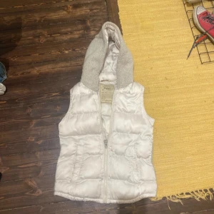 Vit dunväst med huva - Säljer en fluffig vit dunväst från Jofama med dragkedja framtill och en mysig grå huva i mjukt material. Perfekt att slänga över en hoodie eller tröja för extra värme. Västen har en quiltad design och känns riktigt skön att bära.