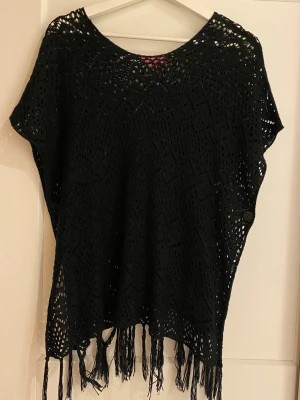 Svart stickad topp med fransar - Säljer en svart stickad topp med korta ärmar och snyggt hålmönster. Toppen har rund hals och fransar längs nederkanten som ger en bohemisk vibe. Perfekt att slänga över ett linne eller bikini på sommaren.