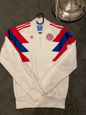 Adidas Fc Bayern München  - Säljer en vit Adidas FC Bayern München jacka med röda och blå detaljer på axlarna. Jackan har dragkedja framtill, ribbade muddar och broderat klubbmärke på bröstet. Materialet är polyester och designen har klassiska Adidas-ränder på axlarna.