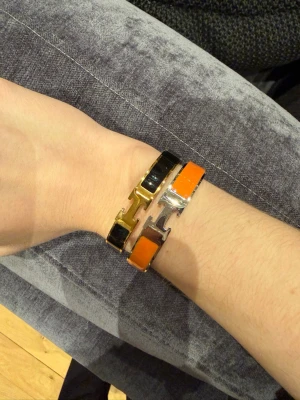 Orange och svart armband  - Två stilrena armband i metall, ett med orange och silverfärgad finish och ett med svart och guldfärgad finish. Båda har den ikoniska H-detaljen framtill och är perfekta för att ge din outfit en lyxig touch. Säljes tillsammans eller separat.