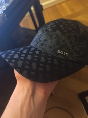 Svart mönstrad keps från BOSS - Snygg svart keps från BOSS med justerbar rem baktill. Kepsen har ett diskret mönster med BOSS-loggor över hela ytan och är tillverkad i ett glansigt syntetmaterial. Perfekt för dig som vill ha en stilren accessoar med lite edge.