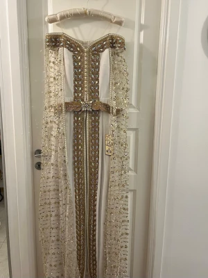 Champagnefärgad långklänning Maher Tex - Exklusiv långklänning i champagnefärg med glittriga paljetter och broderade detaljer i guld och silver. Klänningen har ett vackert bälte med strass och transparenta ärmar dekorerade med paljetter. Materialet är polyester och tyll, perfekt för att sticka ut.