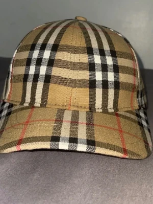Burberry keps - Bilden visar en rutig Burberry keps i beige, svart, vit och röd, inte en väst. Vänligen ladda upp en bild på en väst för att få en relevant beskrivning.