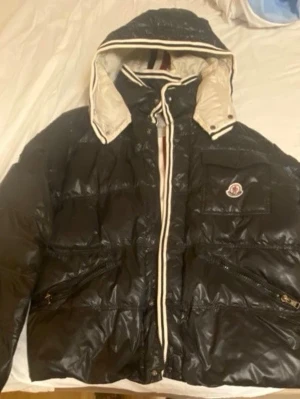 Svart dunjacka från Moncler med huva - Säljer en svart pufferjacka från Moncler med dunfyllning och blank finish. Jackan har vit insida på huvan, dragkedjor och knappar med Moncler-logga, samt praktiska fickor. Perfekt för dig som vill ha en snygg och varm jacka med exklusiv känsla.