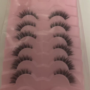 Svarta lösögonfransar i 5-pack - Säljer ett set med fem par svarta lösögonfransar i en rosa plastförpackning. Fransarna är täta och fluffiga med en dramatisk volym och längd, perfekta för att ge ögonen extra intensitet. Materialet är syntetiskt och fransarna har en böjd form.