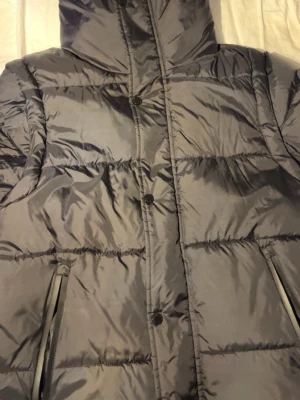 Svart dunjacka från Burberry - Säljer en svart dunjacka/dunväst från Burberry med huva och quiltad design. Västen har två fickor med dragkedja där Burberry-loggan syns på zipparna. Stor Burberry London England-patch på höger arm. Perfekt för kyliga dagar och riktigt snygg stil.