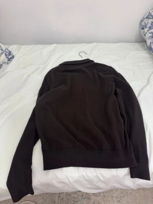 Mörkbrun pikétröja från these glory days - Snygg mörkbrun pikétröja från  these glory days med klassisk krage och knappar framtill. Tröjan är långärmad och har ribbade muddar vid ärmslut och nederkant. Perfekt för dig som gillar stilrena och enkla plagg med en modern touch.