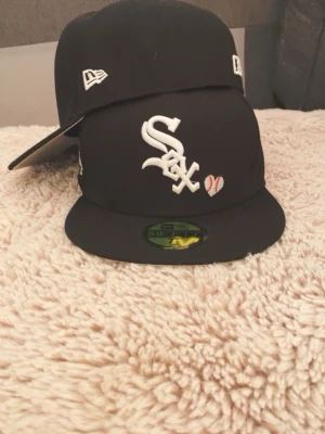 Svart White Sox New Era keps - Säljer en svart New Era 59FIFTY White Sox keps med broderade detaljer som 'Love & Hustle = Game' och 'World Series 05'. Kepsen har vit logga framtill, ett litet basebollhjärta och grå underskärm. Klassisk rak skärm och broderade MLB-loggor på sidorna.
